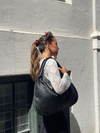 Femme portant un grand sac à main noir tressé, vêtue d'un chemisier blanc en dentelle, posant devant un mur blanc. Mode urbaine élégante.