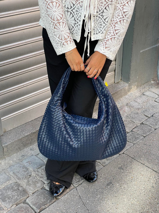 Femme tenant un sac à main bleu tressé, vêtue d'un chemisier en dentelle blanche et pantalon noir, devant une porte métallique. Mode urbaine élégante.