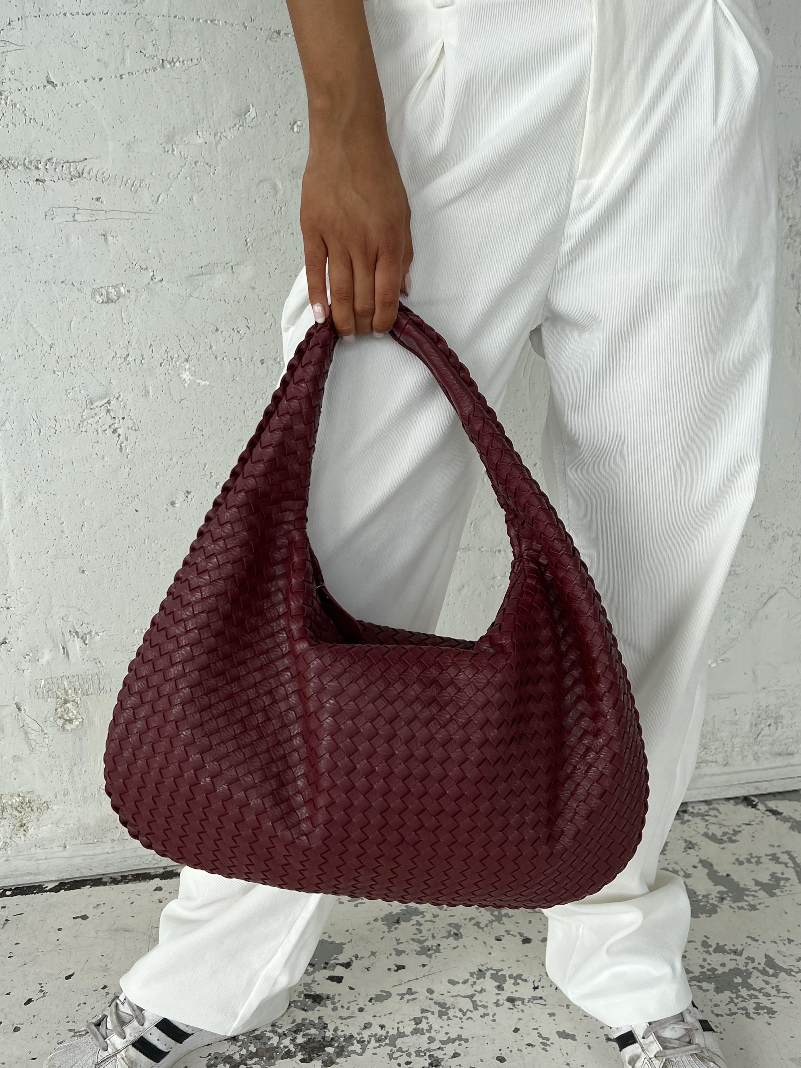 Sac à main tressé bordeaux porté par une personne en pantalon blanc. Accessoire mode tendance, design élégant, idéal pour tenues décontractées.