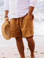 Homme en short en lin marron et chemise blanche, tenant un chapeau de paille sur une plage. Mode estivale, tenue décontractée, bord de mer.