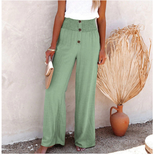 Femme portant un pantalon vert taille haute avec boutons, tenant une pochette beige. Décor minimaliste avec vase en terre cuite et plante séchée.