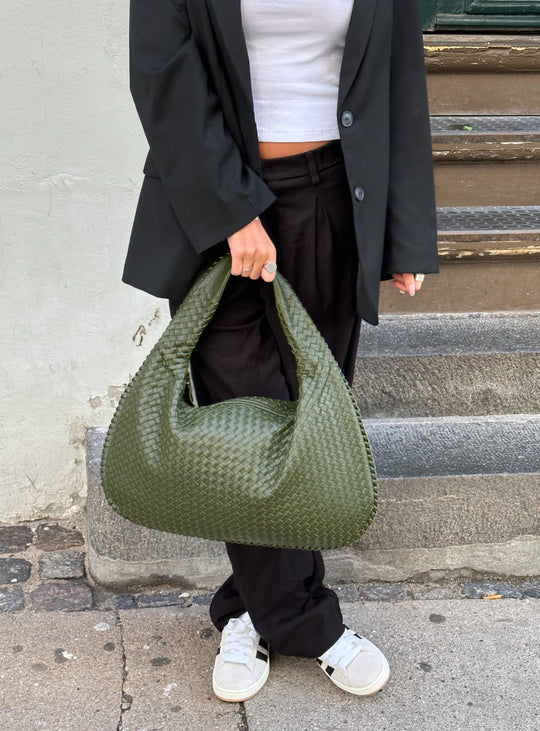 Sac à main vert tissé porté par une personne en tenue noire et baskets blanches, debout sur un trottoir. Mode urbaine, accessoire tendance.
