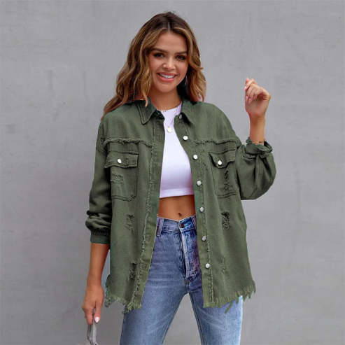 Femme souriante portant une veste en jean vert kaki, chemise blanche et jean bleu, posant devant un mur gris. Mode décontractée, tendance automne.