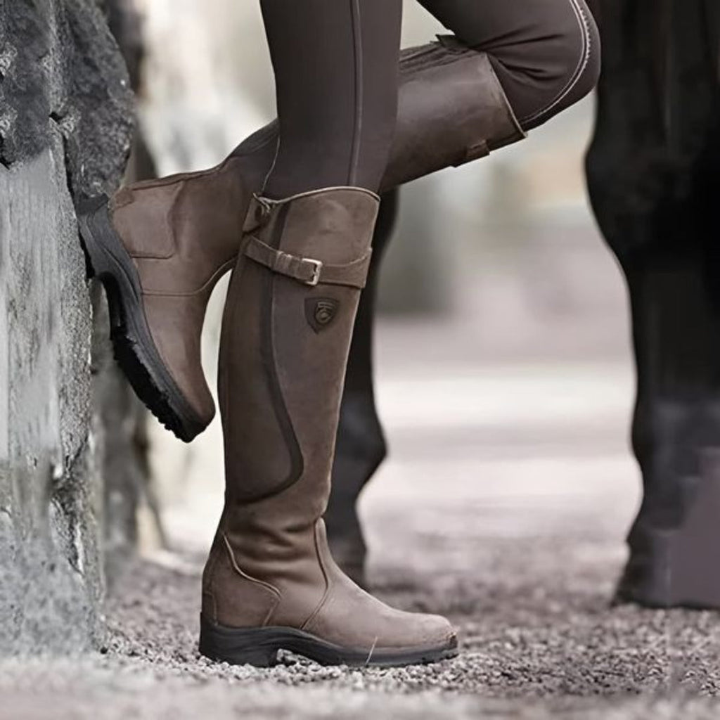 Bottes d'équitation marron en cuir, portées par une personne en pantalon d'équitation, appuyée contre un mur en pierre. Mode équestre élégante.