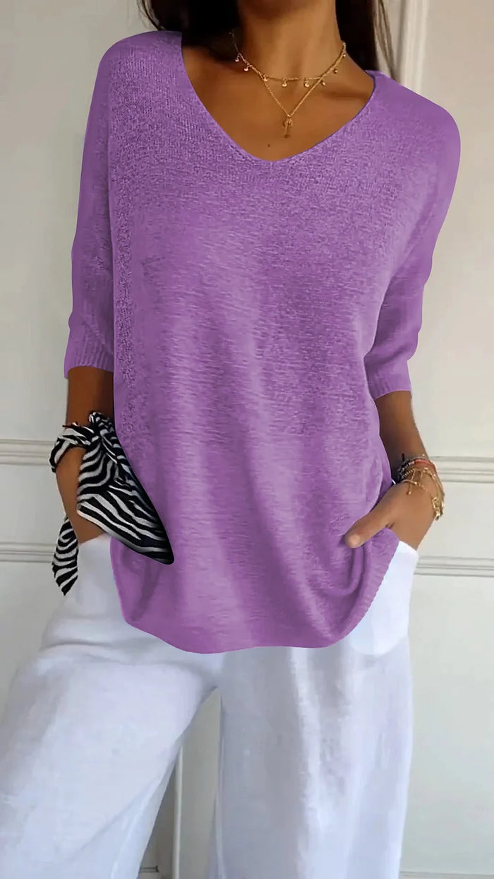Femme portant un pull violet à manches longues et un pantalon blanc, accessoirisé d'un foulard zébré au poignet, devant un mur blanc.