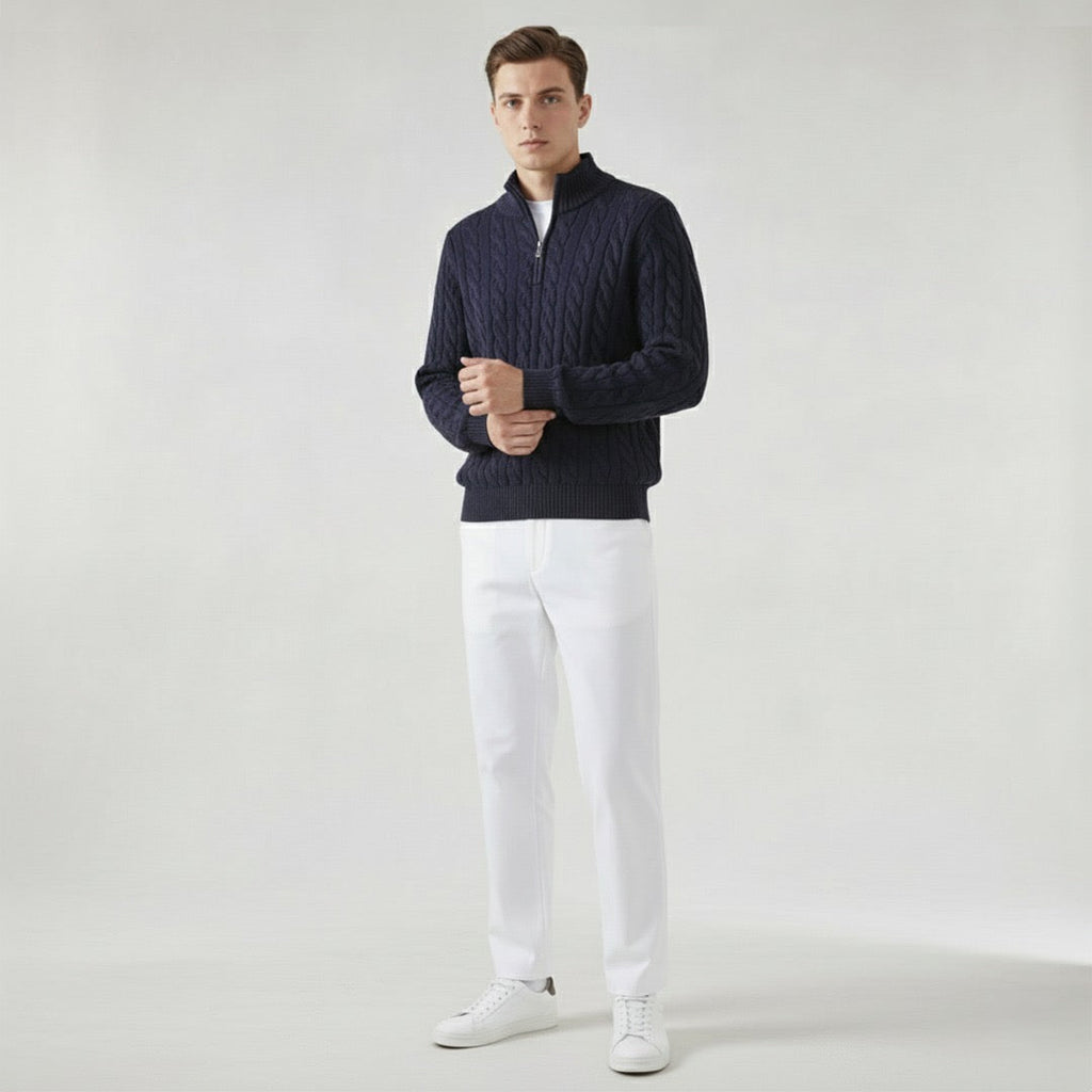 Homme portant un pull bleu marine en maille torsadée, pantalon blanc et baskets blanches, posant sur fond neutre. Mode masculine élégante et moderne.