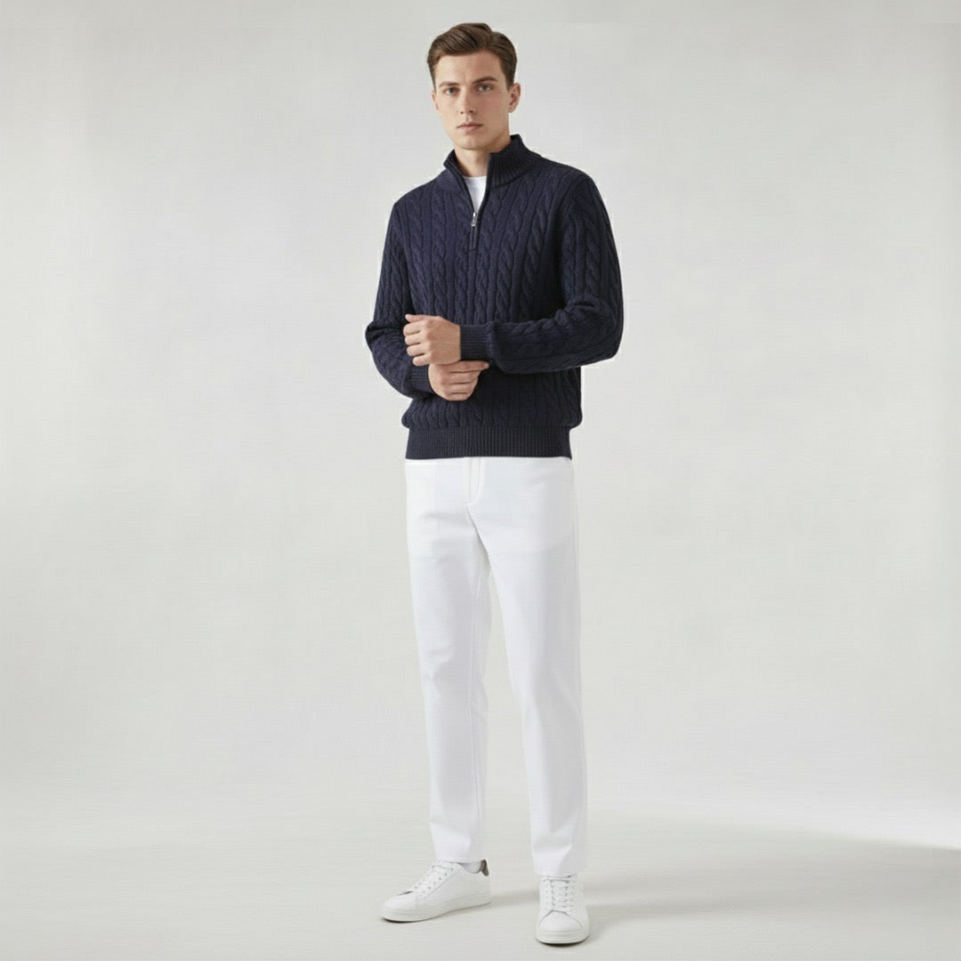 Homme portant un pull bleu marine en maille torsadée, pantalon blanc et baskets blanches, posant sur fond neutre. Mode masculine élégante et moderne.