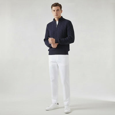 Homme portant un pull bleu marine en maille torsadée, pantalon blanc et baskets blanches, posant sur fond neutre. Mode masculine élégante et moderne.