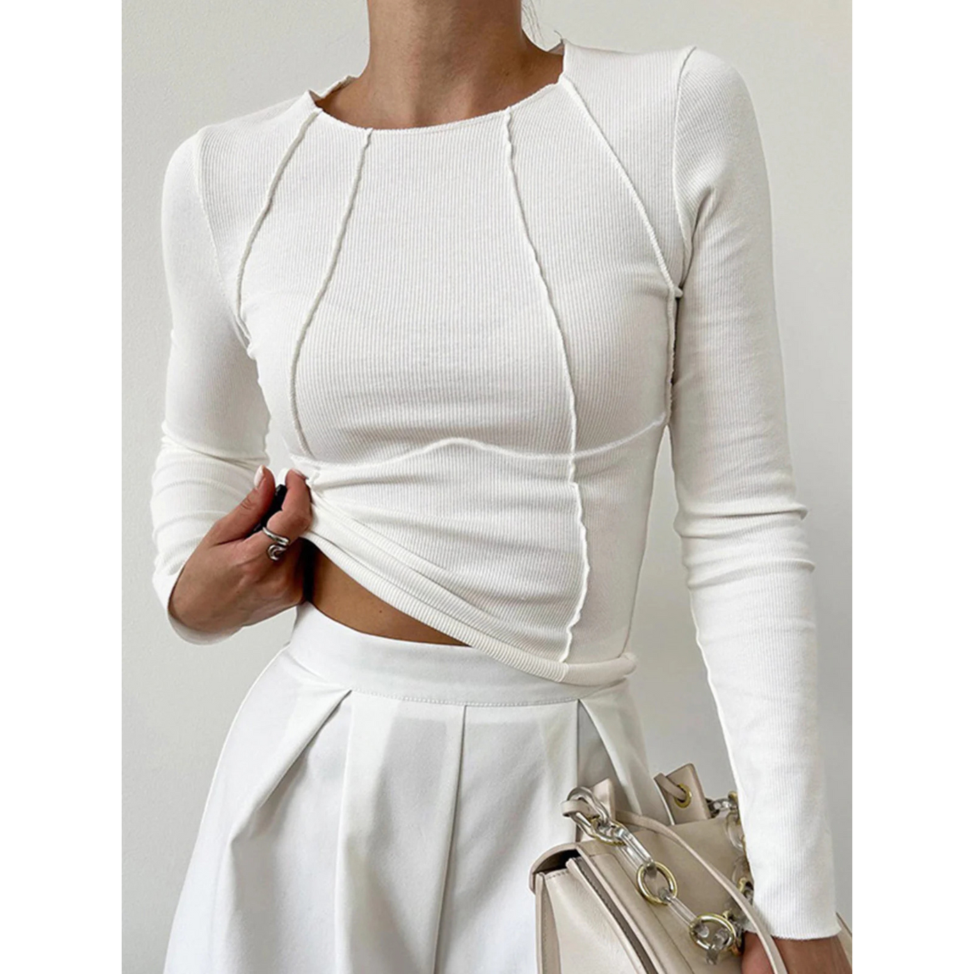 Femme portant un haut blanc ajusté à manches longues et une jupe blanche plissée, tenant un sac à main beige avec chaîne dorée. Mode élégante et minimaliste.