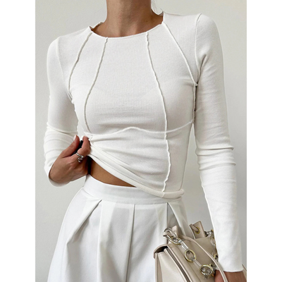 Femme portant un haut blanc ajusté à manches longues et une jupe blanche plissée, tenant un sac à main beige avec chaîne dorée. Mode élégante et minimaliste.