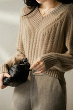 Femme portant un pull en laine beige à col en V, tenant un sac à main tressé noir. Mode automne-hiver, style élégant et décontracté.
