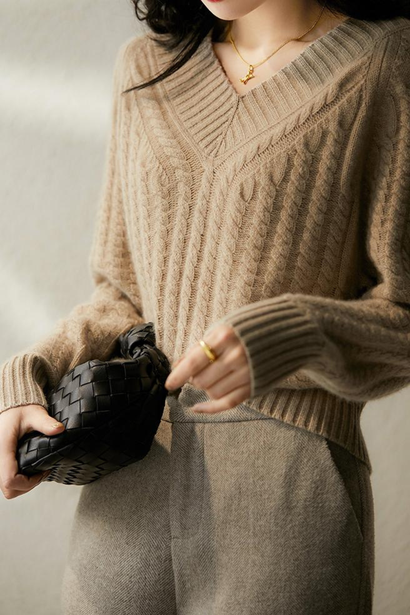 Femme portant un pull en laine beige à col en V, tenant un sac à main tressé noir. Mode automne-hiver, style élégant et décontracté.