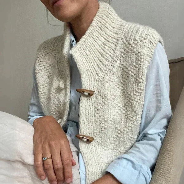 Femme portant un gilet en laine tricoté beige avec boutons en bois, assise sur un canapé. Mode automne, style décontracté, confort élégant.