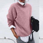 Femme portant un pull rose ample, chemise blanche et pantalon gris, tenant un sac noir. Mode décontractée, tendance automne-hiver.