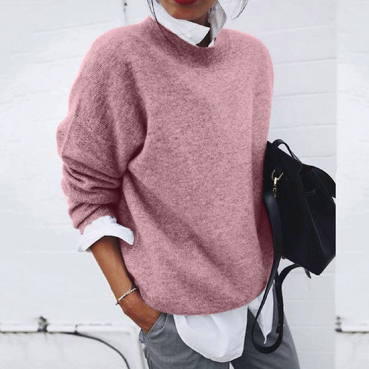Femme portant un pull rose ample, chemise blanche et pantalon gris, tenant un sac noir. Mode décontractée, tendance automne-hiver.