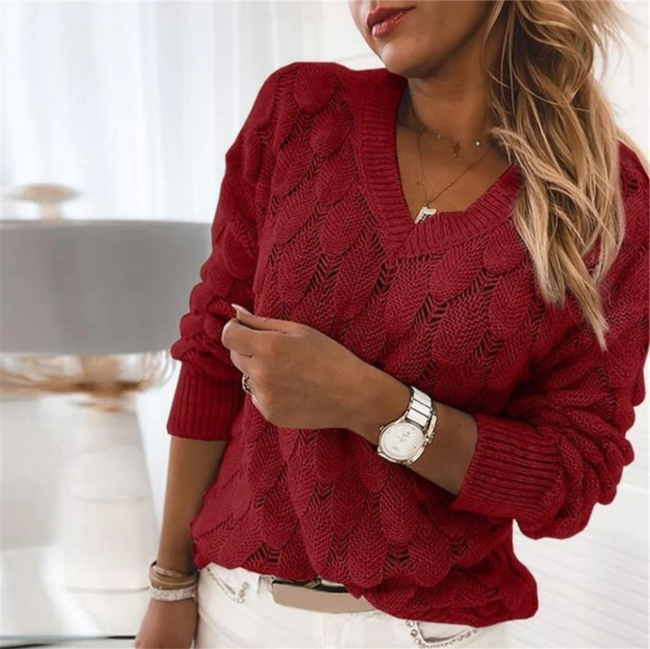 Femme portant un pull rouge en tricot ajouré, montre argentée, cheveux blonds ondulés, intérieur lumineux. Mode automne-hiver élégante.