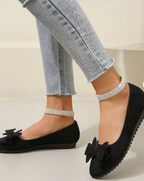 Ballerines noires élégantes avec nœuds, ornées de bracelets de cheville en strass, portées avec un jean slim. Chaussures mode femme chic.