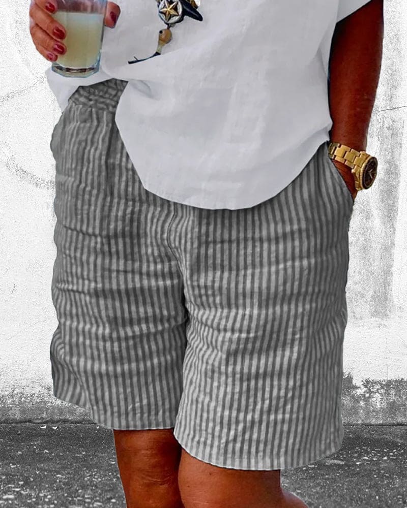 Homme en short rayé gris et chemise blanche, tenant un verre. Montre dorée au poignet. Mode estivale décontractée pour homme.