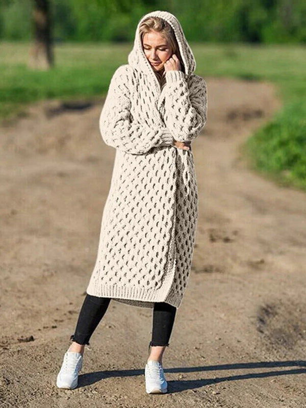 Femme portant un long cardigan en tricot beige avec capuche, debout sur un chemin de terre, style décontracté, mode automne-hiver.