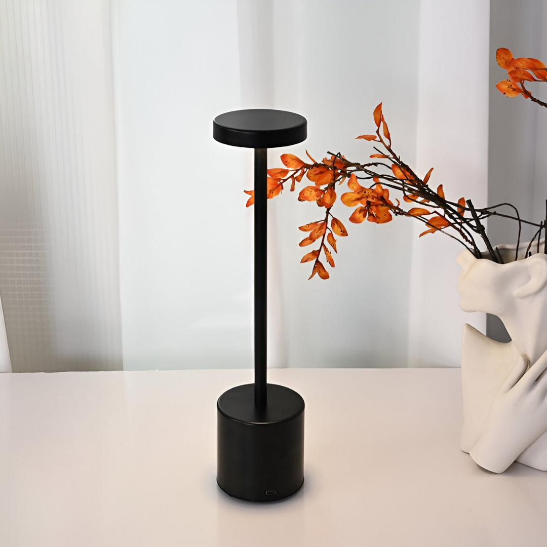 Lampe de table noire moderne sur une table blanche, à côté d'un vase blanc avec des branches orange. Design minimaliste, éclairage intérieur élégant.