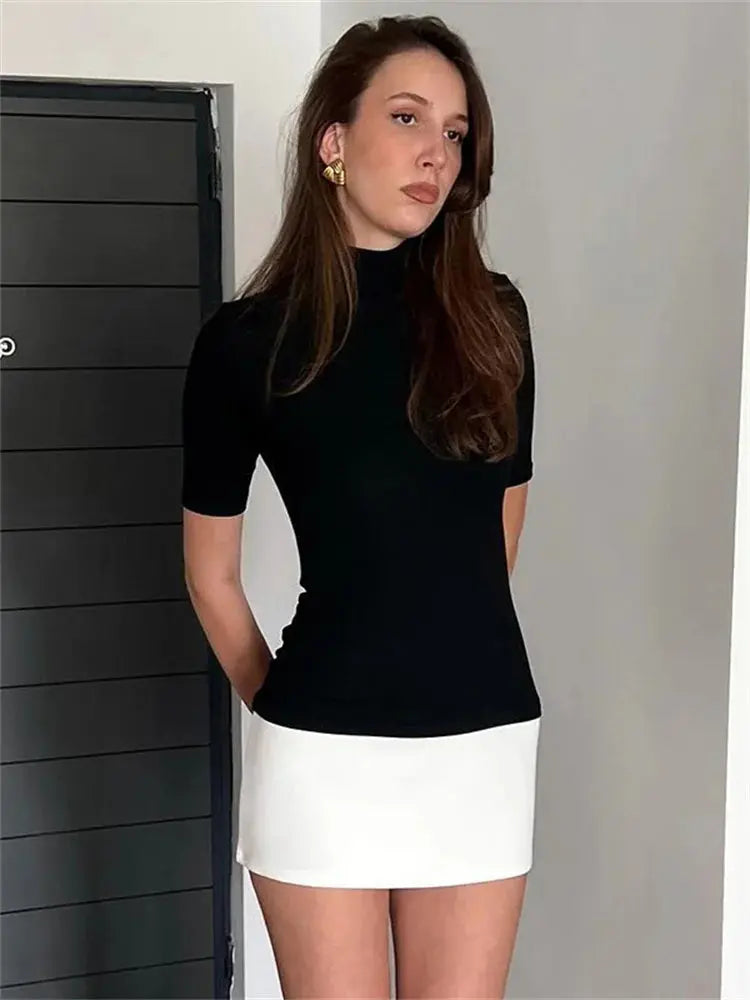 Jeune femme portant un haut noir à manches courtes et une jupe blanche, posant devant une porte noire. Mode élégante, style minimaliste.