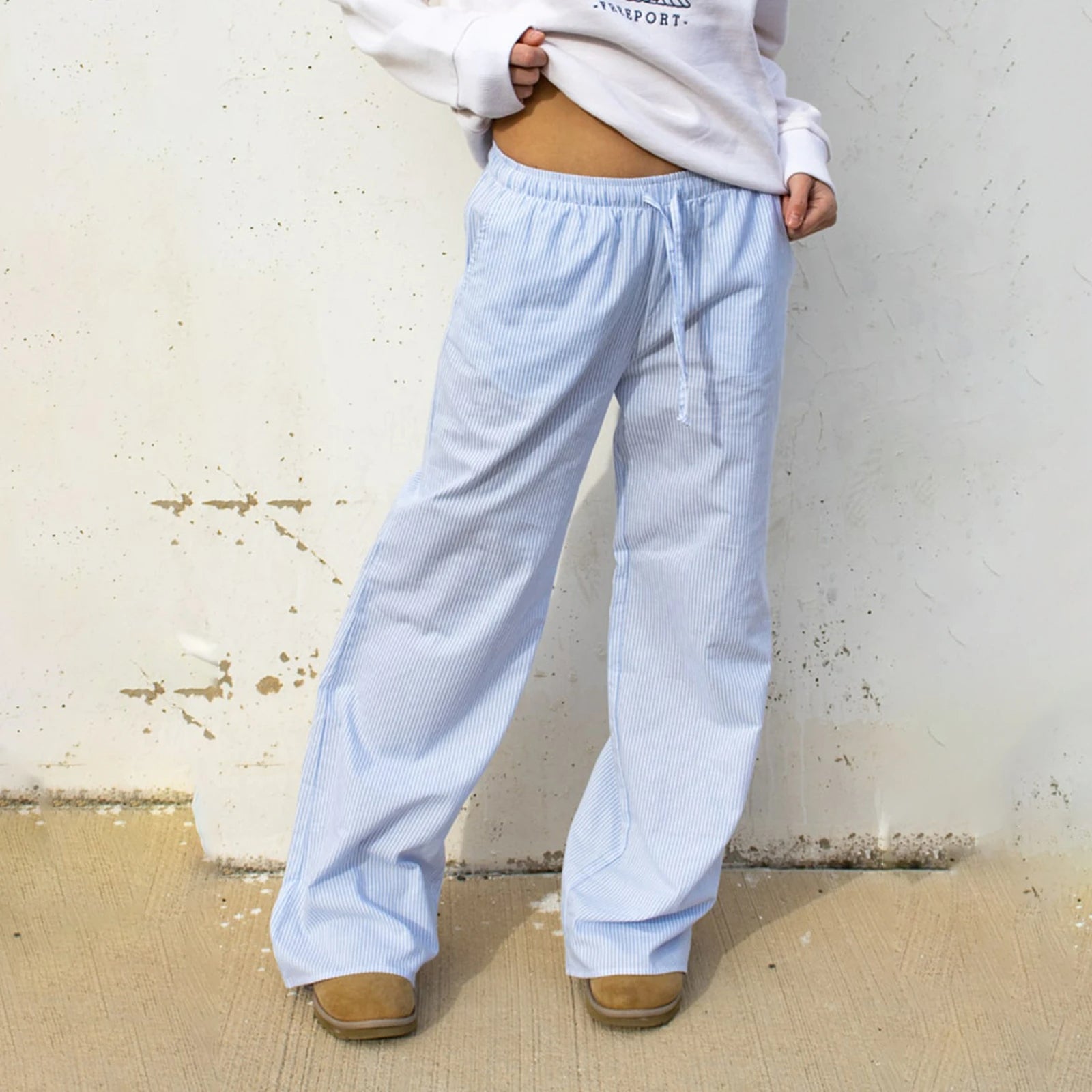 Personne portant un pantalon large rayé bleu et blanc, associé à un sweat-shirt blanc et des bottes marron, posant contre un mur blanc.
