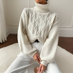 Femme portant un pull torsadé blanc à col roulé, manches longues, assise sur un tapis blanc. Mode hiver élégante et confortable.