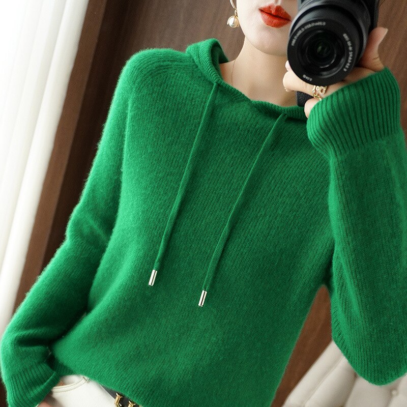 Femme portant un pull vert en laine avec capuche, tenant un appareil photo. Mode automne-hiver, style décontracté, photographie intérieure.