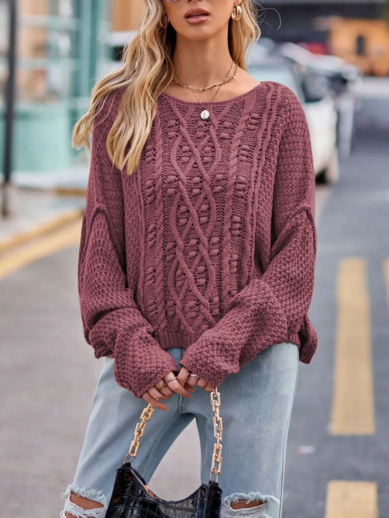 Femme portant un pull en maille torsadée rose, jeans décontractés, tenant un sac à main noir. Mode automne, style urbain chic.