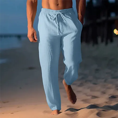 Homme marchant sur la plage au crépuscule, portant un pantalon en lin bleu clair. Mode décontractée, soirée d'été, confort et style.