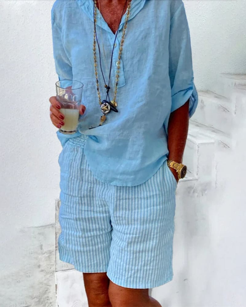 Homme en chemise en lin bleu clair et short rayé, tenant un verre. Style estival décontracté, accessoires dorés, ambiance plage.