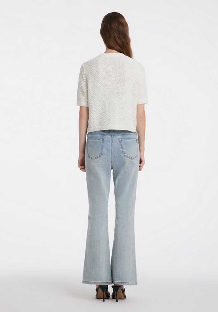 Femme de dos portant un jean évasé bleu clair et un haut blanc texturé, posant sur fond blanc. Mode féminine, style décontracté, tendance 2023.