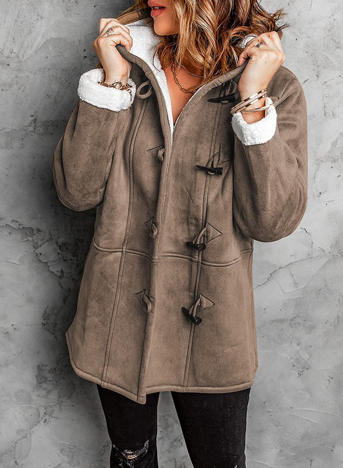 Femme portant un manteau en daim marron avec doublure en fourrure blanche, boutons en bois, posant contre un mur gris. Mode hiver élégante et confortable.