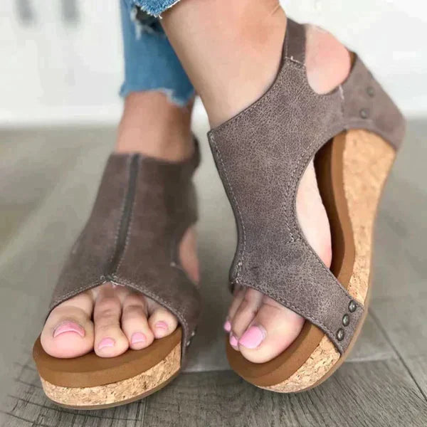 Sandales compensées en liège pour femme, couleur taupe, avec lanières en cuir synthétique, portées sur un sol en bois. Mode estivale confortable.