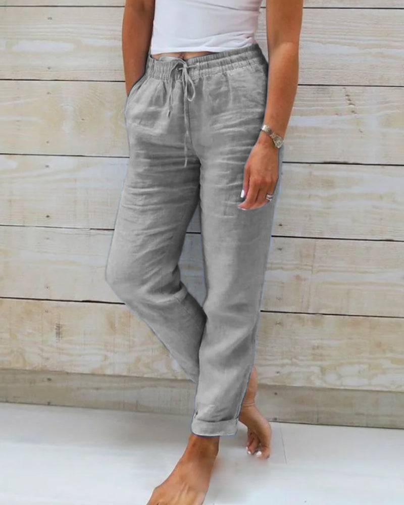 Pantalon en lin gris pour femme, coupe décontractée, avec poches et cordon de serrage, porté avec un haut blanc, sur fond de bois clair.