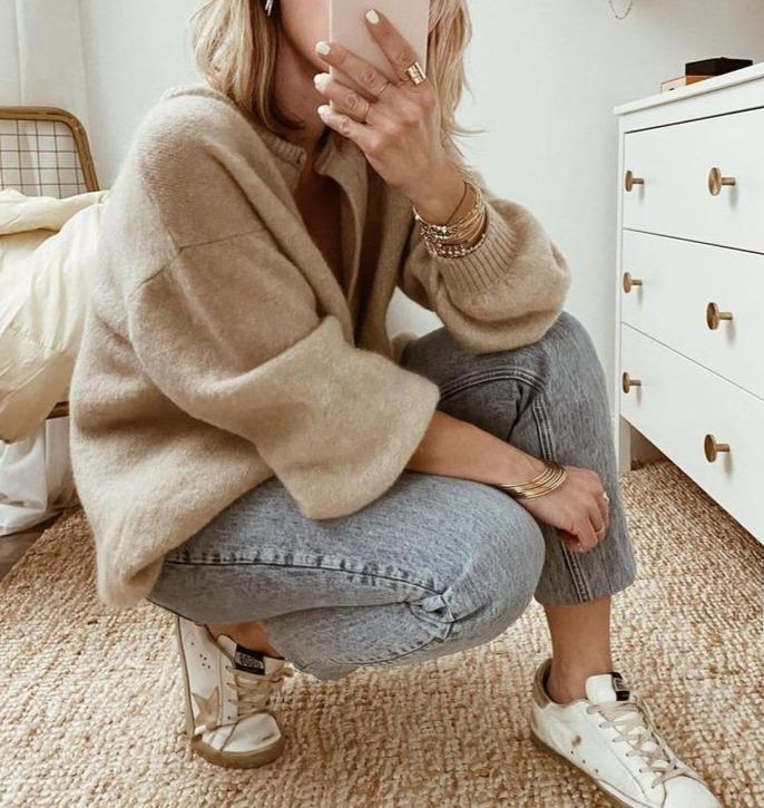Femme en pull beige et jeans, accroupie sur un tapis beige, prenant un selfie. Mode décontractée, baskets blanches, chambre moderne.