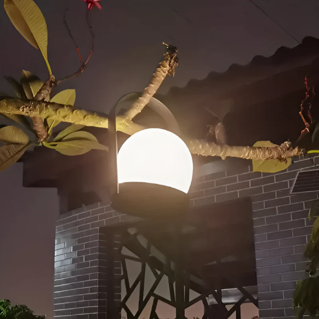 Lampe extérieure moderne accrochée à une branche d'arbre, éclairant une façade en briques noires la nuit. Éclairage jardin, design contemporain.