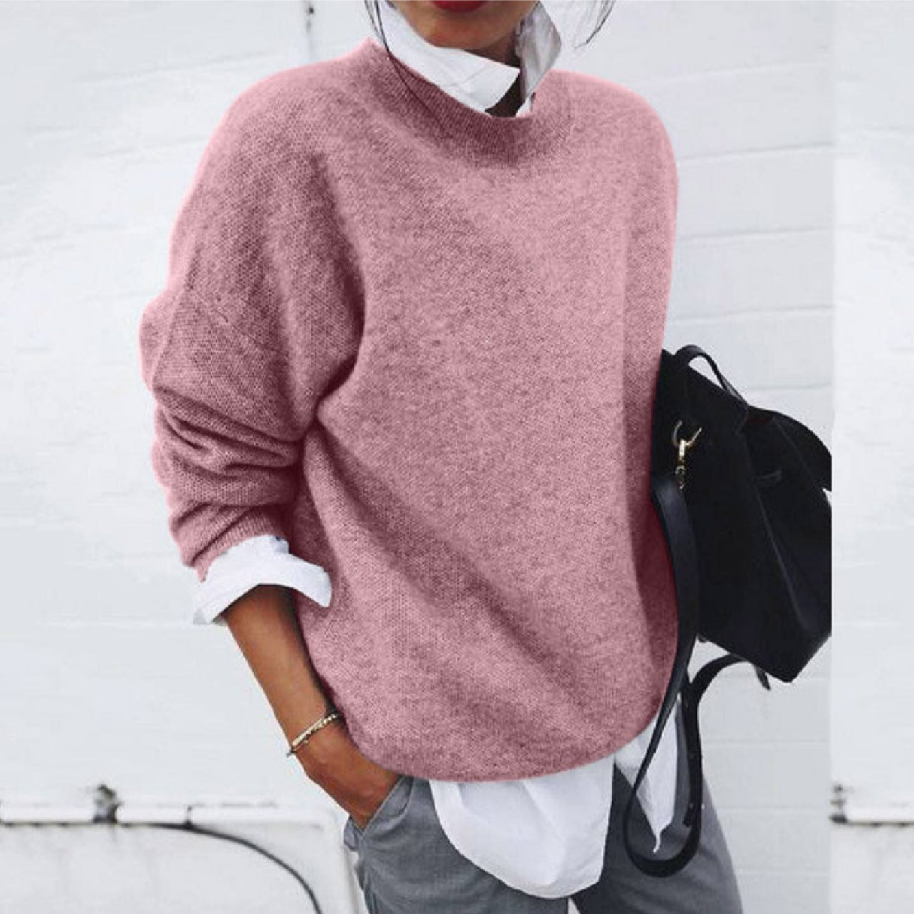 Femme portant un pull rose ample, chemise blanche et pantalon gris, tenant un sac noir. Mode décontractée, tendance automne-hiver.