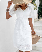 Robe blanche en dentelle brodée, manches courtes, portée par une femme tenant un chapeau de paille et un sac en osier, style estival élégant.