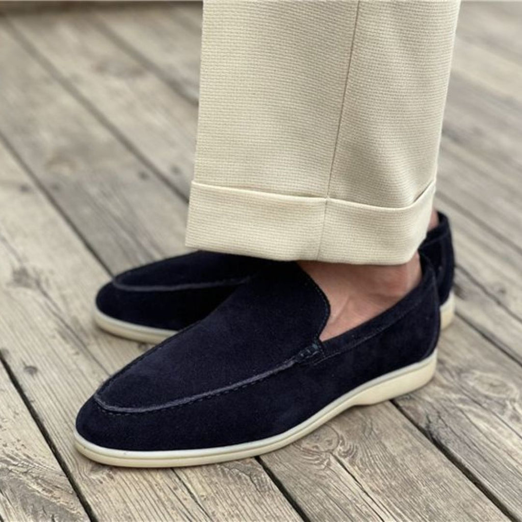 Chaussures mocassins en daim bleu marine pour homme, portées avec un pantalon beige sur une terrasse en bois. Style élégant et décontracté.