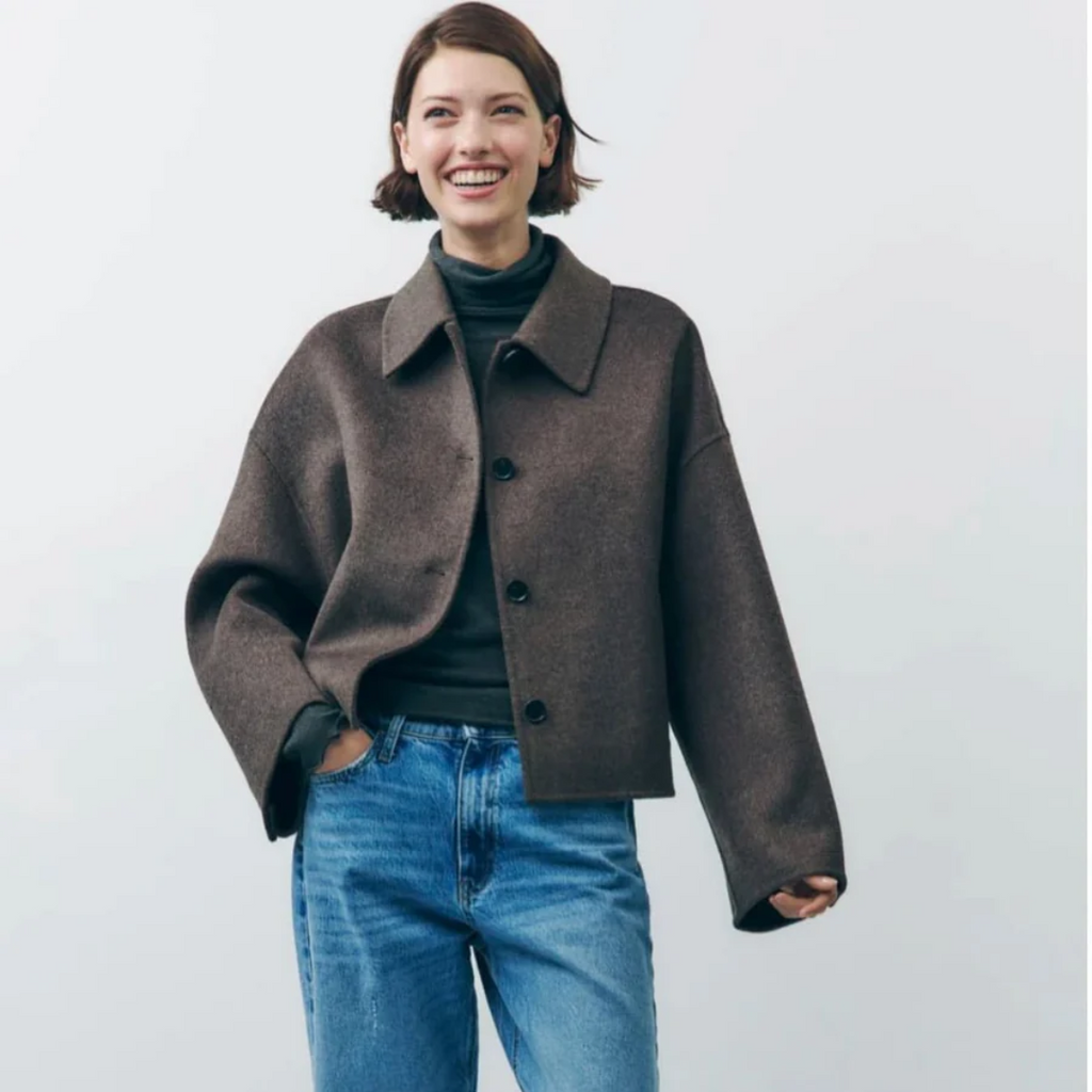 Jeune femme souriante portant une veste marron oversize et un jean bleu, posant devant un fond blanc. Mode automne-hiver, style décontracté.