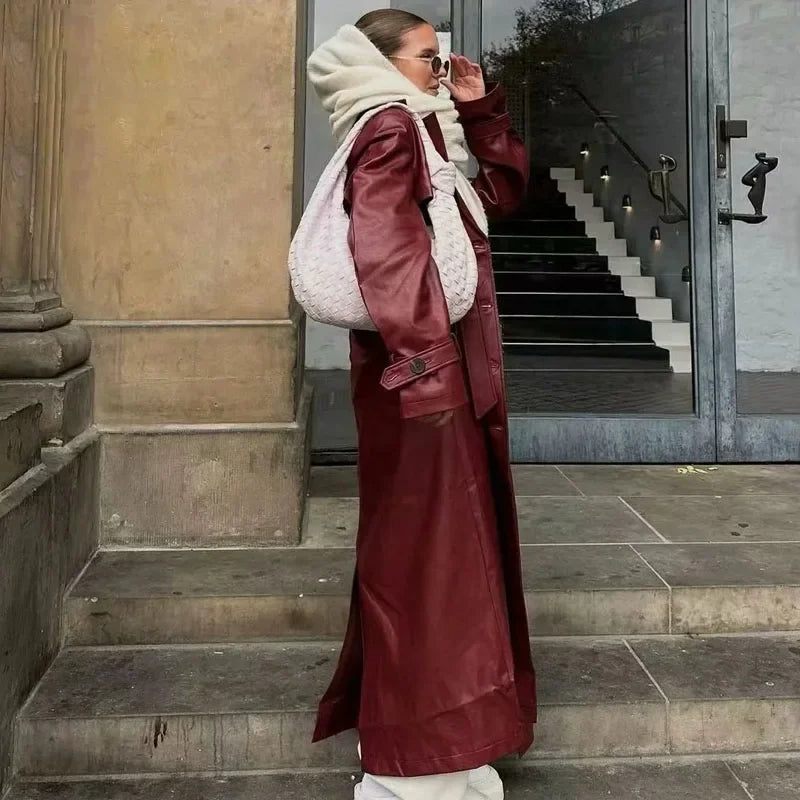 Femme en manteau long en cuir rouge, portant un sac à main blanc et des lunettes de soleil, posant sur des marches devant une porte moderne.