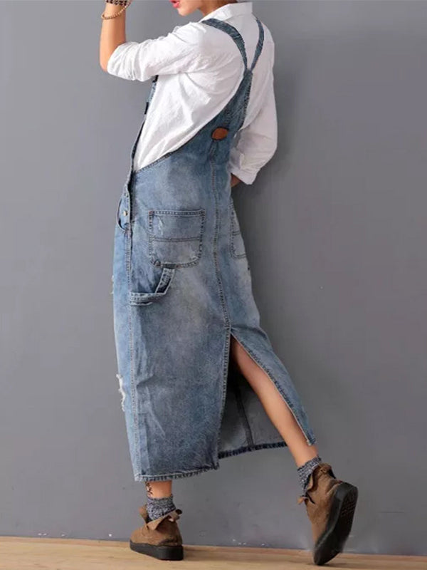 Femme portant une robe salopette en jean avec chemise blanche, posant contre un mur gris. Mode décontractée, tendance denim, style urbain.