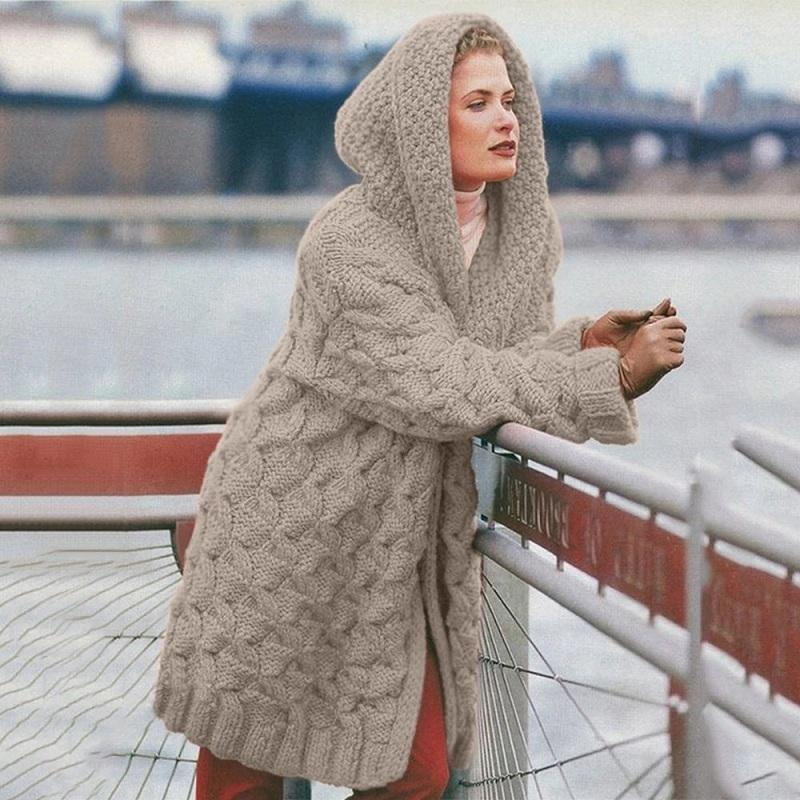 Femme portant un manteau en laine tricoté à capuche, appuyée sur une rambarde près d'un fleuve urbain. Mode hiver, style décontracté, paysage urbain.