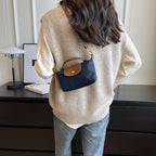 Femme de dos portant un pull beige, chemise blanche et sac à main bleu marine. Mode automne, style décontracté, intérieur moderne.