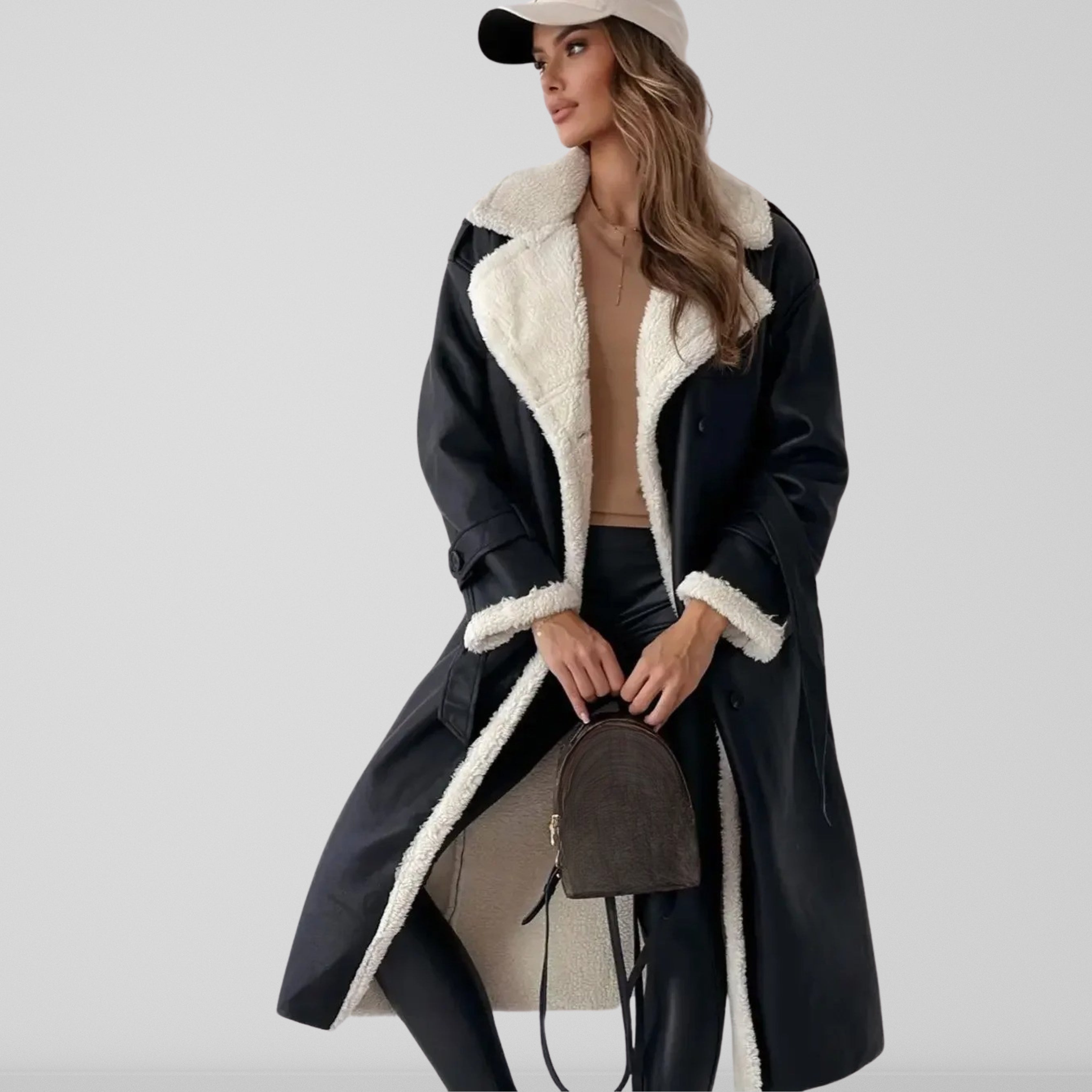 Femme portant un manteau long en cuir noir avec doublure en fourrure blanche, tenue chic avec pantalon noir, casquette beige et sac à dos marron.