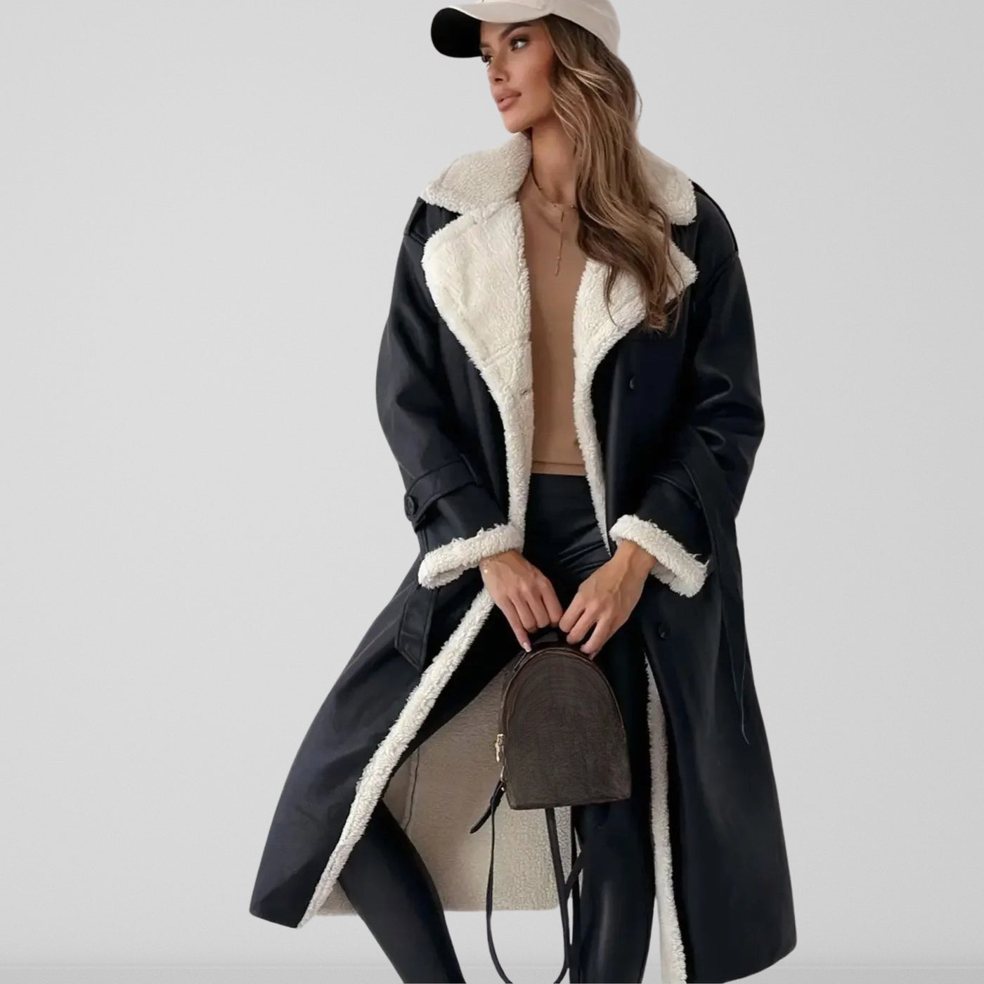 Femme portant un manteau long en cuir noir avec doublure en fourrure blanche, tenue chic avec pantalon noir, casquette beige et sac à dos marron.