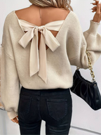 Femme portant un pull beige avec un grand nœud dans le dos, tenant un sac à main noir. Mode automne, style élégant et décontracté.