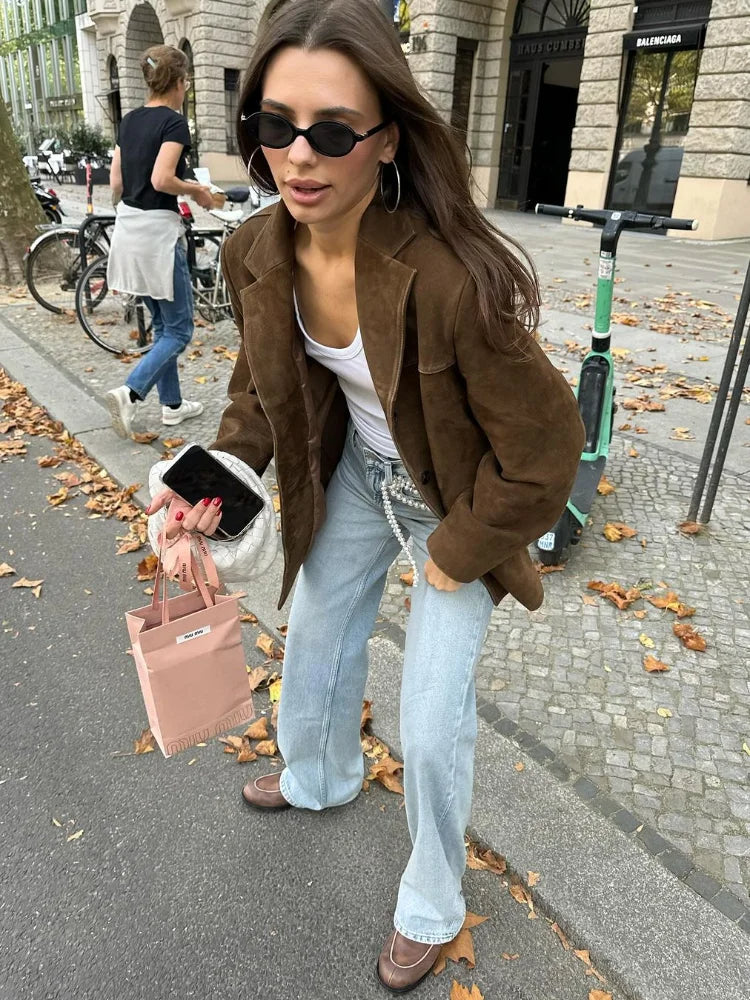 Femme en veste marron, lunettes de soleil, jeans larges, tenant un sac rose et un smartphone, marchant sur un trottoir avec feuilles d'automne.