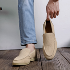 Personne tenant un mocassin beige en daim, portant un jean bleu clair, sur un sol en bois. Chaussure élégante, mode décontractée, style moderne.
